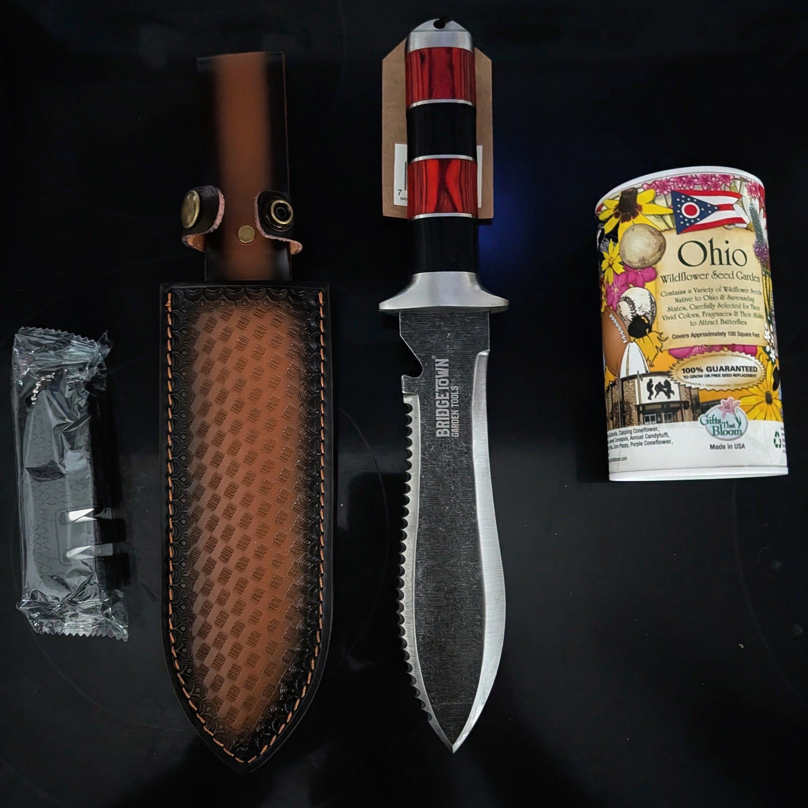 A fixed-blade knife with a serrated silver blade and a red-and-black wrapped handle, displayed with a brown textured sheath, a small wrapped accessory, and a printed card on a black background.
