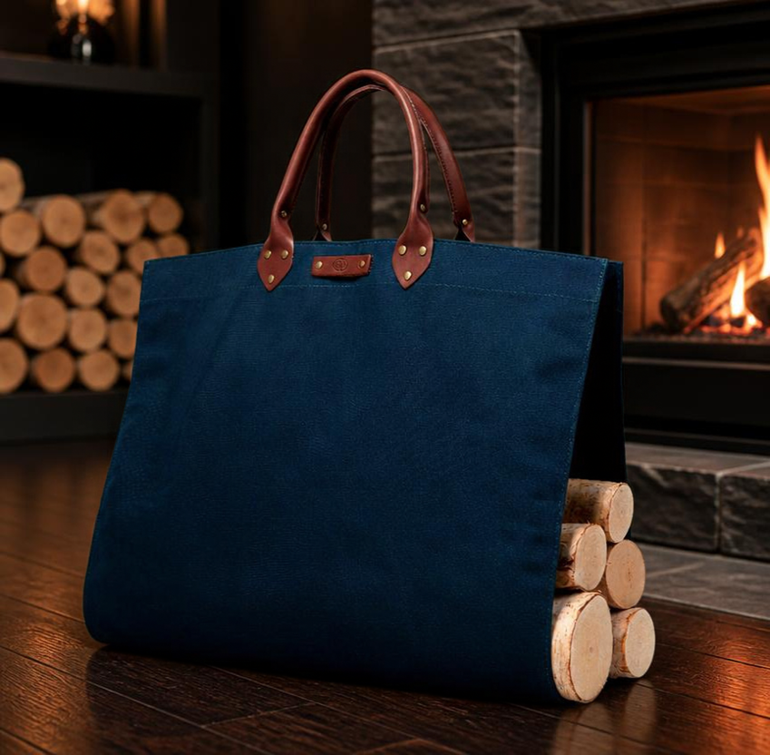 Navy blue canvas log carrier with leather handles loaded with firewood on dark hardwood floor next to a modern fireplace