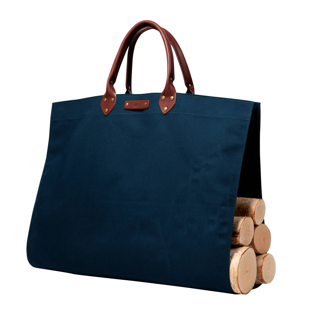 A navy blue canvas tote bag with black leather handles and brass grommets, shown holding a stack of wooden logs on a plain white background.